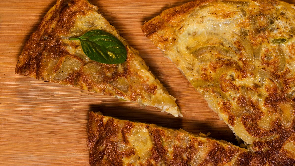 Tortilla spagnola tagliata a spicchi su un tagliere di legno, con patate e uova